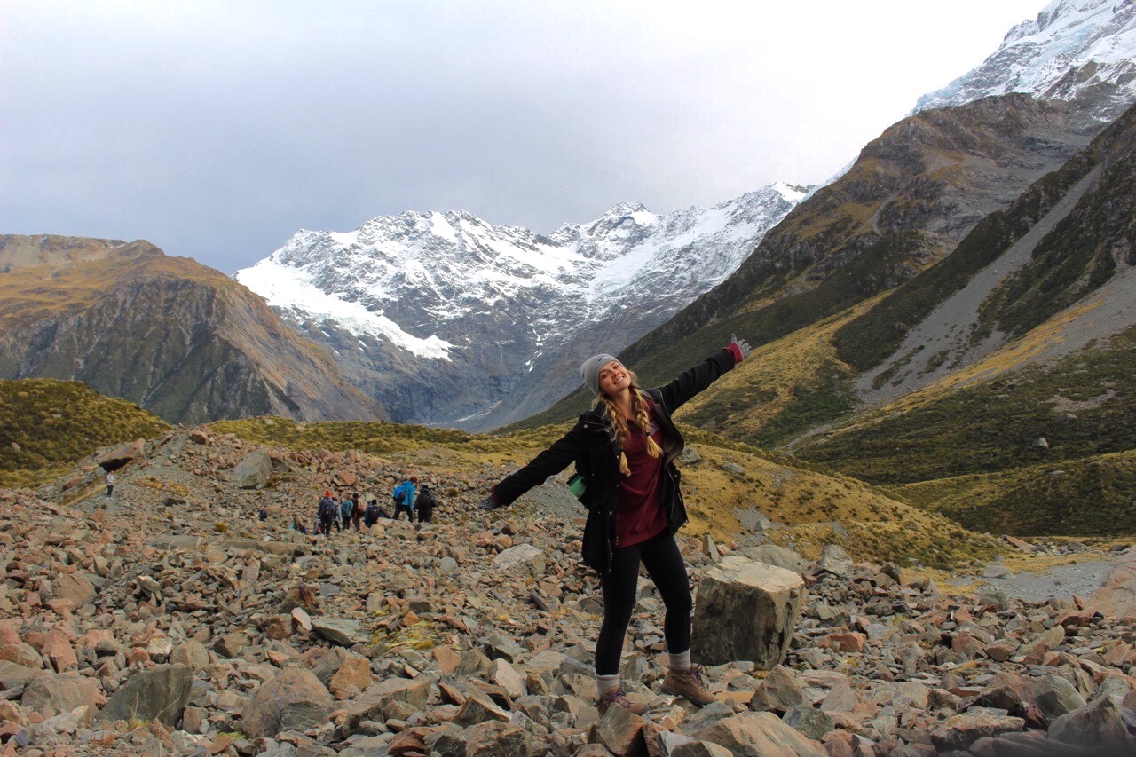 Girl stands with arms raised in front of Mount Cook mountain range.