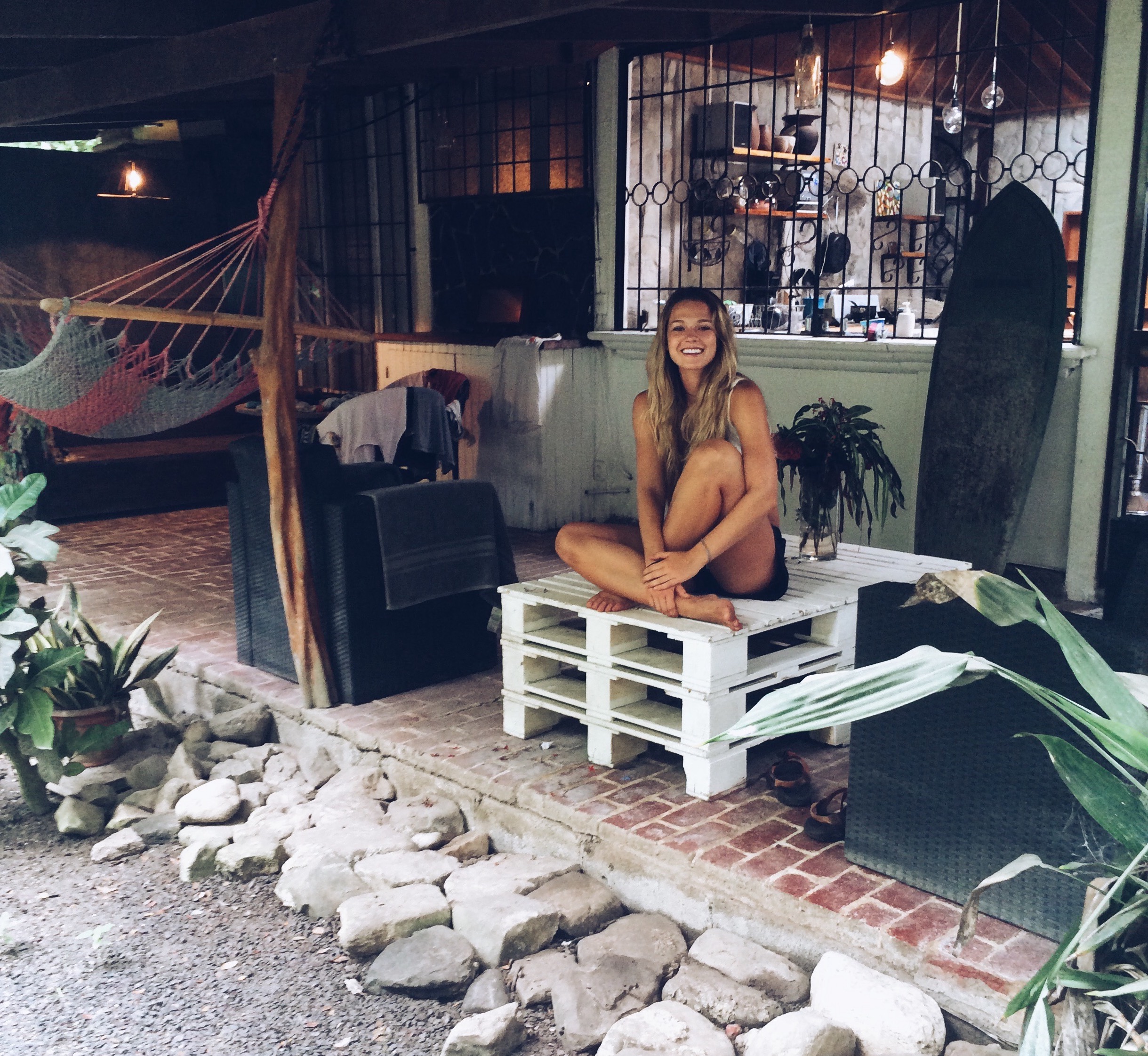 Girl relaxes on a porch with a surfboard and hammock behind her.